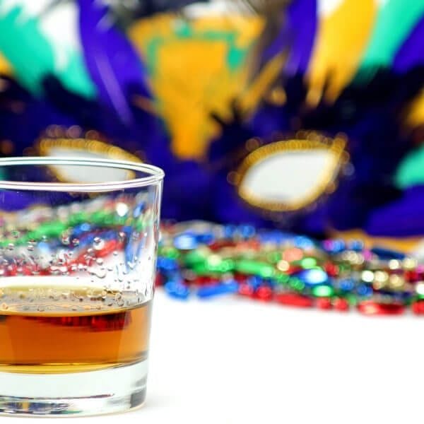 Bourbon in a glass in front of a feathered Mardi Gras mask and beads.